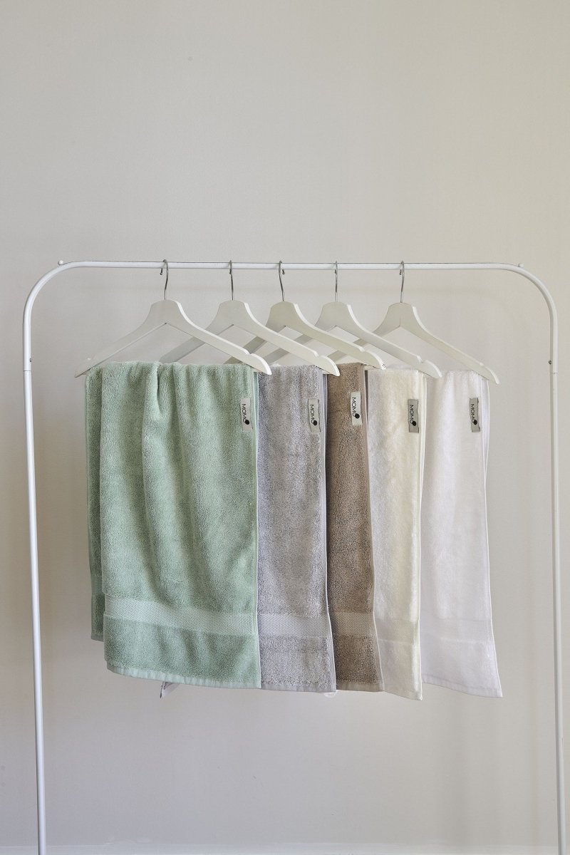 Vijf pastelkleurige bamboe handdoeken op witte houten hangers tegen neutrale achtergrond in groen, grijs, bruin, crème en