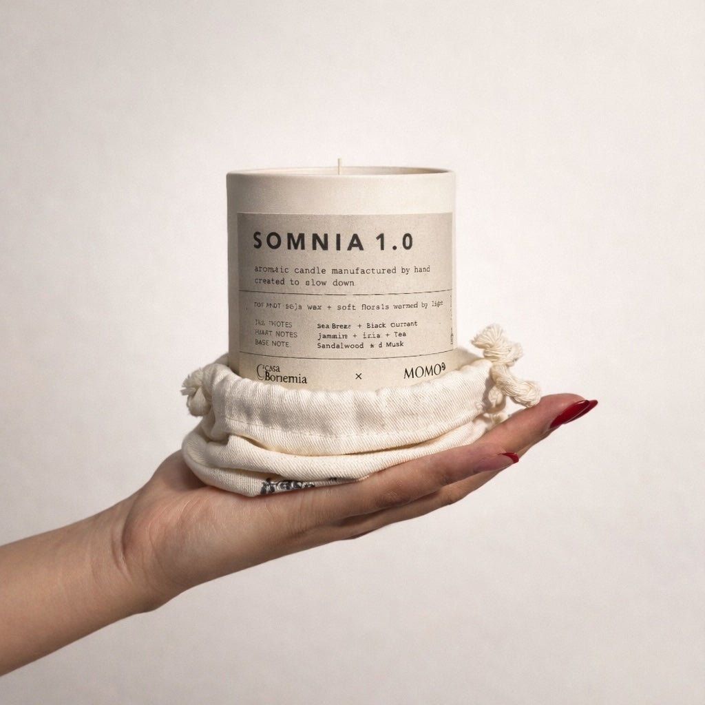 Hand holding cream-colored Somnia 1.0 aromatic candle wrapped in natural cotton fabric against white background