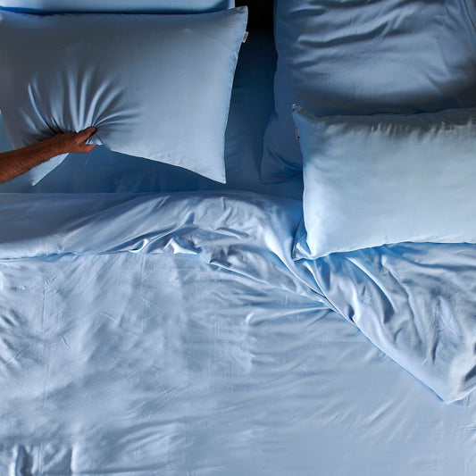 Pessoa arrumando jogo de cama de bambu azul claro com almofadas na cama, mostrando textura e maciez do jogo de roupa completo