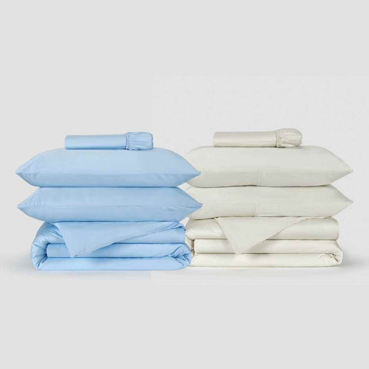 Twee Momo Cotton dekbedovertreksets naast elkaar, één lichtblauw en één crème, met gevouwen lakens en opgerolde bedlinnen