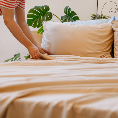 Pessoa a arranjar lençol e fronha de bambú cor de pêssego na cama com plantas monstera ao fundo